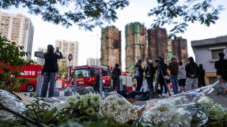 Flores no chão aparecem em primeiro plano com os prédios incendiados ao fundo