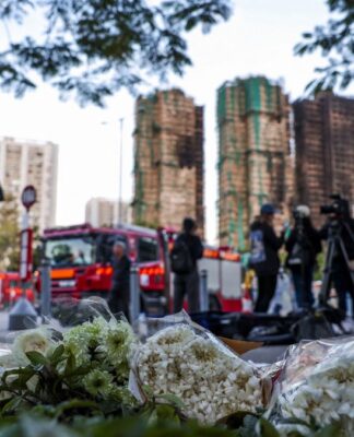 Flores no chão aparecem em primeiro plano com os prédios incendiados ao fundo