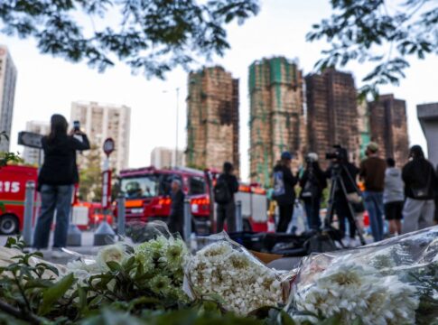 Flores no chão aparecem em primeiro plano com os prédios incendiados ao fundo