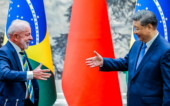 Dois líderes políticos, um do Brasil e outro da China, sorriem e estendem as mãos para um cumprimento diante das bandeiras de seus países.