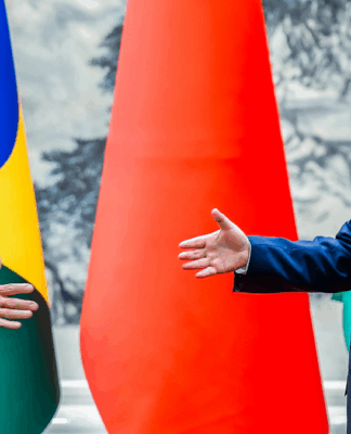 Dois líderes políticos, um do Brasil e outro da China, sorriem e estendem as mãos para um cumprimento diante das bandeiras de seus países.