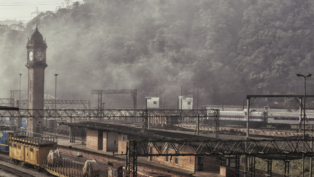 Paisagem da estação ferroviária de Paranapiacaba, com torre do relógio em destaque e trilhos ao redor. A cena está envolta por névoa densa, com vagões e trens visíveis diante de uma encosta coberta por mata.