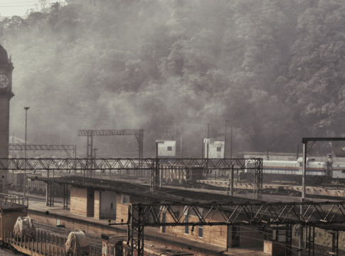 Paisagem da estação ferroviária de Paranapiacaba, com torre do relógio em destaque e trilhos ao redor. A cena está envolta por névoa densa, com vagões e trens visíveis diante de uma encosta coberta por mata.