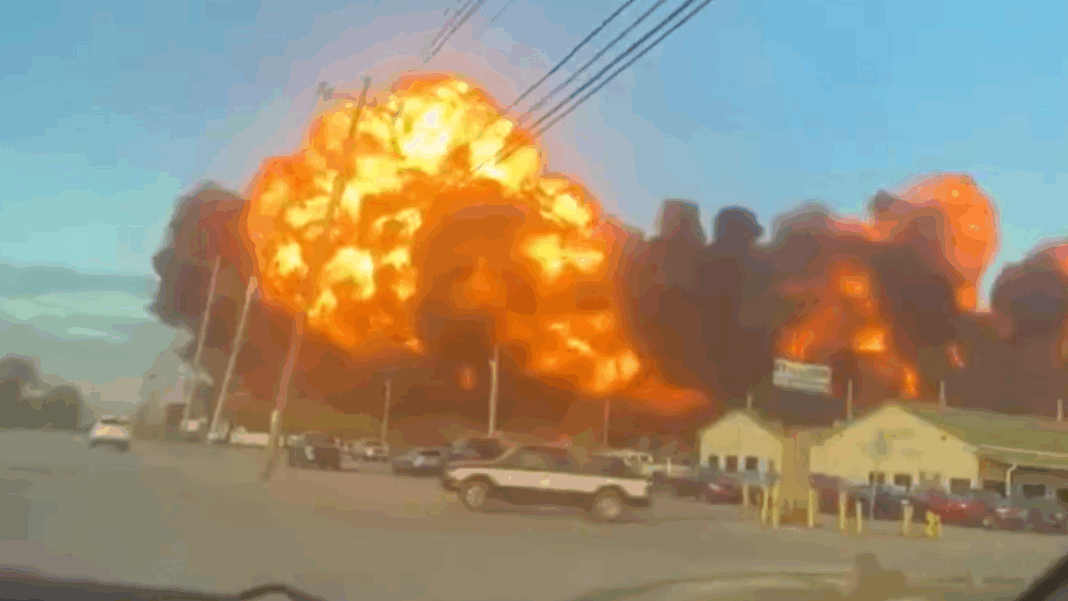 Grande explosão em área industrial, com uma bola de fogo laranja e densa fumaça preta se espalhando pelo céu, enquanto carros estão estacionados e algumas construções aparecem em primeiro plano.