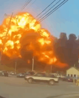 Grande explosão em área industrial, com uma bola de fogo laranja e densa fumaça preta se espalhando pelo céu, enquanto carros estão estacionados e algumas construções aparecem em primeiro plano.