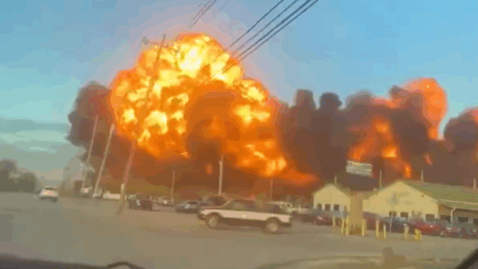 Grande explosão em área industrial, com uma bola de fogo laranja e densa fumaça preta se espalhando pelo céu, enquanto carros estão estacionados e algumas construções aparecem em primeiro plano.