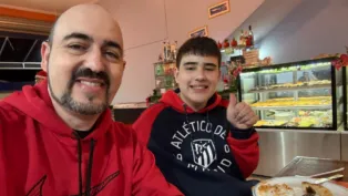 Dois homens sorridentes sentados em uma mesa de restaurante posam para a foto; ambos usam moletom vermelho, um deles com o logotipo da Nike e o outro do Atlético de Madrid, enquanto fazem sinal de positivo com o polegar. Um prato com restos de pizza está à frente, e ao fundo há um balcão com vitrine de salgados e doces.