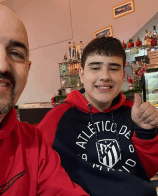 Dois homens sorridentes sentados em uma mesa de restaurante posam para a foto; ambos usam moletom vermelho, um deles com o logotipo da Nike e o outro do Atlético de Madrid, enquanto fazem sinal de positivo com o polegar. Um prato com restos de pizza está à frente, e ao fundo há um balcão com vitrine de salgados e doces.