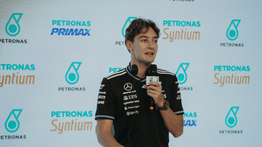 Homem veste camiseta preta da equipe Mercedes-AMG Petronas e segura um microfone durante entrevista ou coletiva de imprensa, com logotipos da Petronas e Syntium exibidos em um painel de fundo.