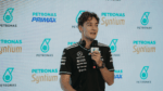 Homem veste camiseta preta da equipe Mercedes-AMG Petronas e segura um microfone durante entrevista ou coletiva de imprensa, com logotipos da Petronas e Syntium exibidos em um painel de fundo.