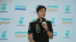 Homem veste camiseta preta da equipe Mercedes-AMG Petronas e segura um microfone durante entrevista ou coletiva de imprensa, com logotipos da Petronas e Syntium exibidos em um painel de fundo.