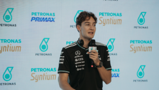 Homem veste camiseta preta da equipe Mercedes-AMG Petronas e segura um microfone durante entrevista ou coletiva de imprensa, com logotipos da Petronas e Syntium exibidos em um painel de fundo.