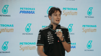 Homem veste camiseta preta da equipe Mercedes-AMG Petronas e segura um microfone durante entrevista ou coletiva de imprensa, com logotipos da Petronas e Syntium exibidos em um painel de fundo.