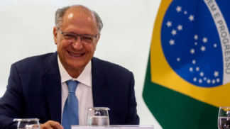 Homem de terno azul e gravata clara sorri sentado à mesa durante uma reunião oficial; à direita aparece parcialmente a bandeira do Brasil ao fundo, junto a copos de água e documentos sobre a mesa.