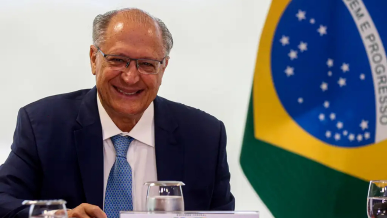 Homem de terno azul e gravata clara sorri sentado à mesa durante uma reunião oficial; à direita aparece parcialmente a bandeira do Brasil ao fundo, junto a copos de água e documentos sobre a mesa.