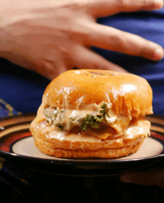 Pessoa sentada segura um prato com um hambúrguer grande e suculento; a mão repousa sobre a barriga coberta por camiseta azul, enquanto o sanduíche aparece em destaque à frente.