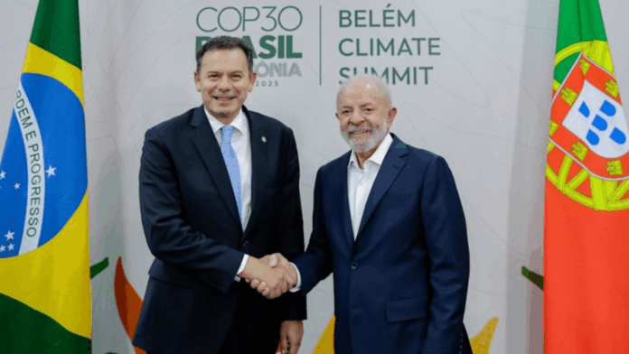 Dois homens de terno sorriem e se cumprimentam com um aperto de mão diante de bandeiras do Brasil e de Portugal, com o painel ao fundo exibindo o texto “COP30 Brasil Amazônia 2025 – Belém Climate Summit”.