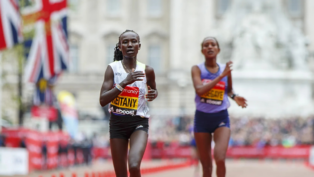 Duas atletas correm próximas à linha de chegada em uma maratona de rua. A corredora à frente, usando camiseta branca e tênis rosa, exibe expressão de esforço. Bandeiras do Reino Unido e o público aparecem ao fundo.