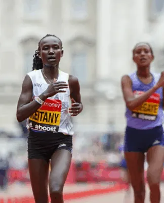 Duas atletas correm próximas à linha de chegada em uma maratona de rua. A corredora à frente, usando camiseta branca e tênis rosa, exibe expressão de esforço. Bandeiras do Reino Unido e o público aparecem ao fundo.