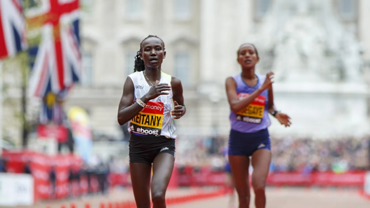 Duas atletas correm próximas à linha de chegada em uma maratona de rua. A corredora à frente, usando camiseta branca e tênis rosa, exibe expressão de esforço. Bandeiras do Reino Unido e o público aparecem ao fundo.