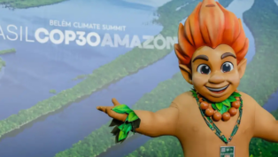 Mascote de aparência infantil, com cabelo laranja em forma de chamas e adereços de folhas verdes, posa sorridente com os braços abertos diante de painel com imagem aérea da Floresta Amazônica e o texto “Brasil COP30 Amazônia – Belém Climate Summit”.