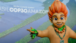 Mascote de aparência infantil, com cabelo laranja em forma de chamas e adereços de folhas verdes, posa sorridente com os braços abertos diante de painel com imagem aérea da Floresta Amazônica e o texto “Brasil COP30 Amazônia – Belém Climate Summit”.