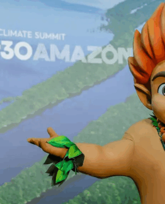 Mascote de aparência infantil, com cabelo laranja em forma de chamas e adereços de folhas verdes, posa sorridente com os braços abertos diante de painel com imagem aérea da Floresta Amazônica e o texto “Brasil COP30 Amazônia – Belém Climate Summit”.