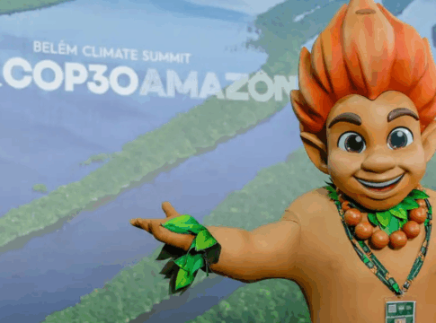 Mascote de aparência infantil, com cabelo laranja em forma de chamas e adereços de folhas verdes, posa sorridente com os braços abertos diante de painel com imagem aérea da Floresta Amazônica e o texto “Brasil COP30 Amazônia – Belém Climate Summit”.