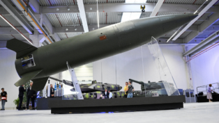 Míssil de grande porte exposto em um suporte transparente dentro de um pavilhão industrial, com tanques militares ao fundo e visitantes observando os equipamentos.
