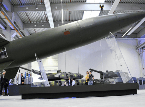 Míssil de grande porte exposto em um suporte transparente dentro de um pavilhão industrial, com tanques militares ao fundo e visitantes observando os equipamentos.
