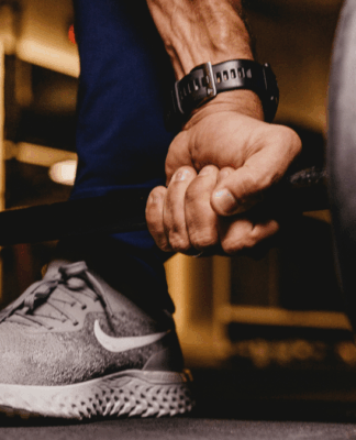 Mão masculina firme segurando uma barra de peso carregada, com foco nos músculos do antebraço e no tênis esportivo cinza, enquanto o ambiente de academia aparece desfocado ao fundo.