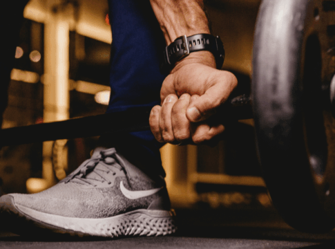 Mão masculina firme segurando uma barra de peso carregada, com foco nos músculos do antebraço e no tênis esportivo cinza, enquanto o ambiente de academia aparece desfocado ao fundo.