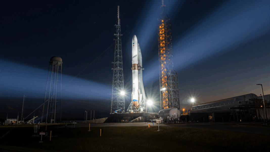 Foguete em plataforma de lançamento iluminado à noite, com fortes refletores formando feixes de luz no céu escuro, enquanto instalações do centro espacial aparecem ao fundo próximo ao horizonte alaranjado.