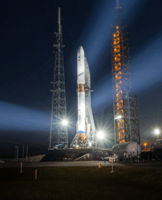 Foguete em plataforma de lançamento iluminado à noite, com fortes refletores formando feixes de luz no céu escuro, enquanto instalações do centro espacial aparecem ao fundo próximo ao horizonte alaranjado.