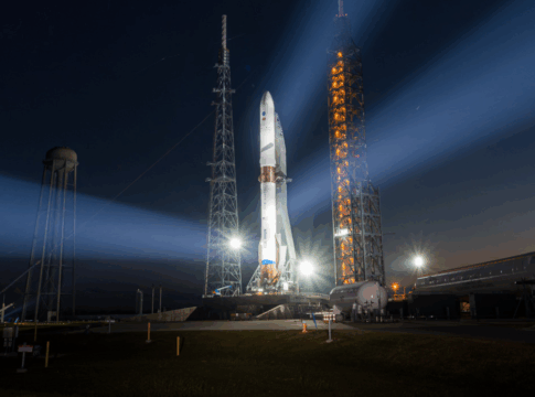 Foguete em plataforma de lançamento iluminado à noite, com fortes refletores formando feixes de luz no céu escuro, enquanto instalações do centro espacial aparecem ao fundo próximo ao horizonte alaranjado.