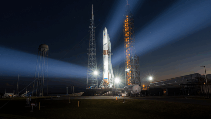 Foguete em plataforma de lançamento iluminado à noite, com fortes refletores formando feixes de luz no céu escuro, enquanto instalações do centro espacial aparecem ao fundo próximo ao horizonte alaranjado.