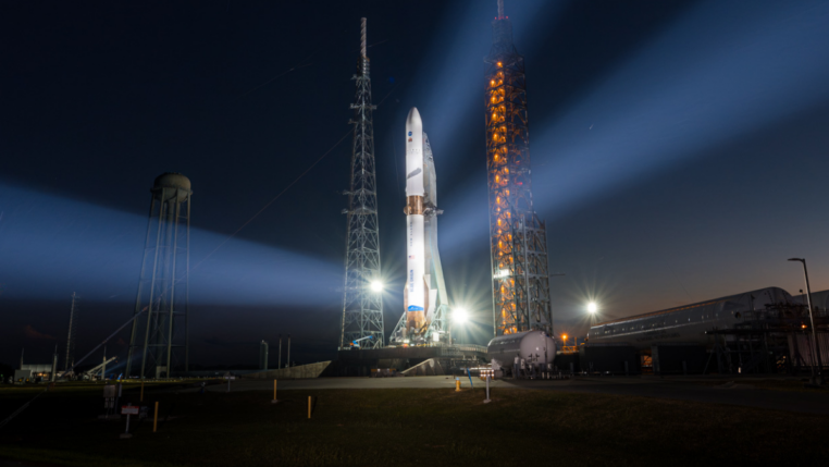 Foguete em plataforma de lançamento iluminado à noite, com fortes refletores formando feixes de luz no céu escuro, enquanto instalações do centro espacial aparecem ao fundo próximo ao horizonte alaranjado.