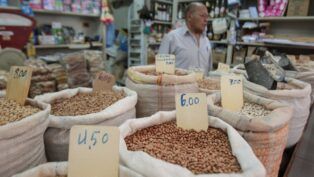 A imagem mostra uma banca de feira, com sacos de graõs e preços variados