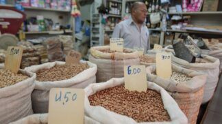 A imagem mostra uma banca de feira, com sacos de graõs e preços variados