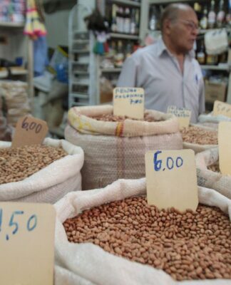 A imagem mostra uma banca de feira, com sacos de graõs e preços variados