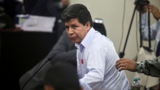 Homem de camisa social clara olha para o lado enquanto se levanta durante uma sessão em ambiente oficial; ao fundo, há câmeras, documentos empilhados e outras pessoas, enquanto uma mão toca seu braço.