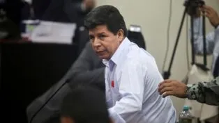 Homem de camisa social clara olha para o lado enquanto se levanta durante uma sessão em ambiente oficial; ao fundo, há câmeras, documentos empilhados e outras pessoas, enquanto uma mão toca seu braço.