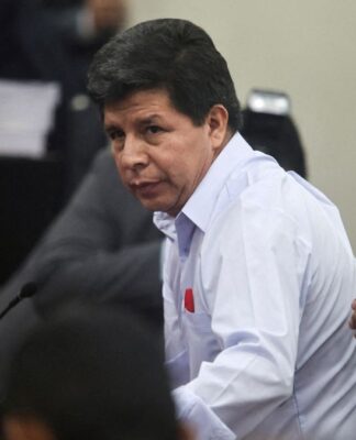 Homem de camisa social clara olha para o lado enquanto se levanta durante uma sessão em ambiente oficial; ao fundo, há câmeras, documentos empilhados e outras pessoas, enquanto uma mão toca seu braço.