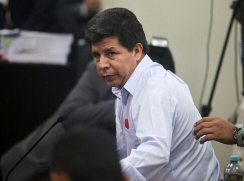 Homem de camisa social clara olha para o lado enquanto se levanta durante uma sessão em ambiente oficial; ao fundo, há câmeras, documentos empilhados e outras pessoas, enquanto uma mão toca seu braço.