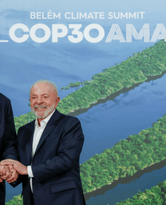 Dois homens de terno sorriem e apertam as mãos diante de um painel com imagem aérea da Floresta Amazônica e o texto “Brasil COP30 Amazônia – Belém Climate Summit”.