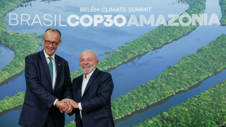 Dois homens de terno sorriem e apertam as mãos diante de um painel com imagem aérea da Floresta Amazônica e o texto “Brasil COP30 Amazônia – Belém Climate Summit”.