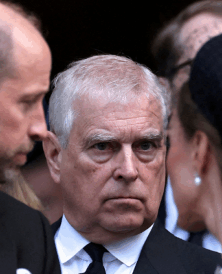 Homem de expressão séria, vestindo terno preto e gravata, é visto entre duas pessoas que conversam próximas a ele durante um evento formal