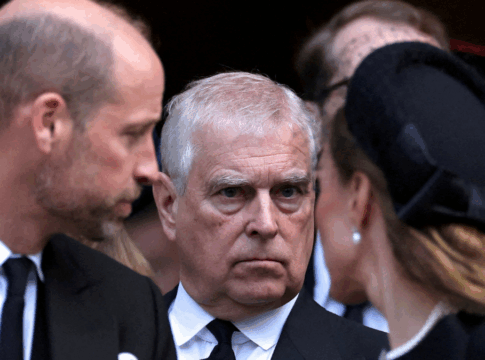 Homem de expressão séria, vestindo terno preto e gravata, é visto entre duas pessoas que conversam próximas a ele durante um evento formal