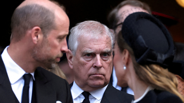 Homem de expressão séria, vestindo terno preto e gravata, é visto entre duas pessoas que conversam próximas a ele durante um evento formal