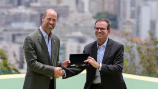 Dois homens de terno trocam um aperto de mão enquanto um deles segura uma caixa aberta com uma chave dourada em seu interior, com prédios desfocados ao fundo.